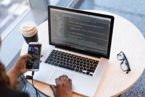 A hand holding a smartphone is next to a laptop displaying coding, with a coffee cup and glasses on a wooden table.