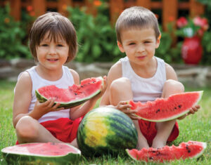 Two children sitting on grass, each holding a slice of watermelon, with a whole watermelon and additional slices around them. They are smiling, wearing tank tops and shorts.