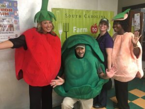 Four people are dressed in colorful fruit and vegetable costumes: a strawberry, a leafy green, a peach, and a tomato, posing joyfully in front of a banner that reads "South Carolina Farm to."