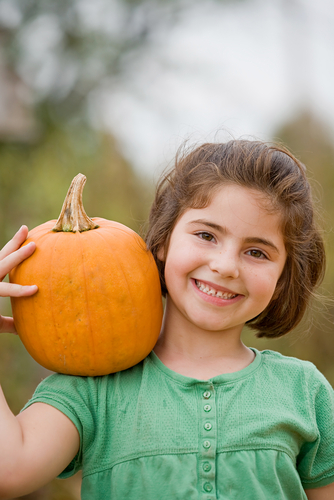 A smiling girl with brown hair holds a pumpkin on her shoulder while wearing a green shirt.