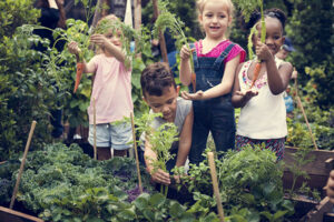 A group of children in a garden, joyfully holding up freshly harvested vegetables. They are surrounded by lush green plants and raised garden beds.