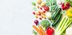A variety of fresh vegetables arranged on a light surface, including potatoes, tomatoes, zucchini, broccoli, carrots, red and yellow bell peppers, garlic, radishes, and leafy greens.