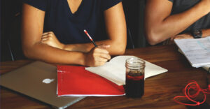 A person's arms resting on a table, one holding a pencil over an open notebook, with a red folder, a laptop, a glass of drink, and headphones visible nearby.
