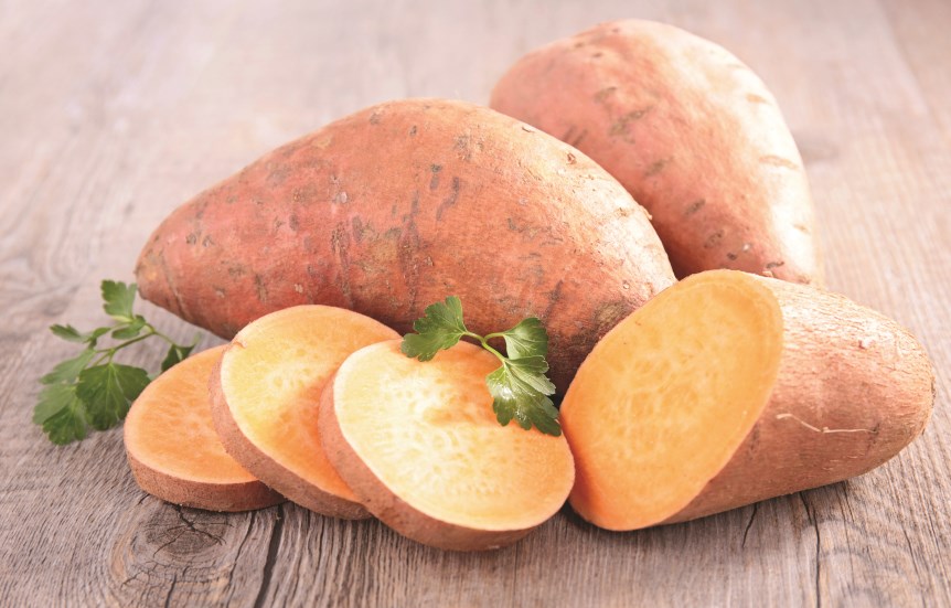A group of whole and sliced orange-fleshed sweet potatoes on a wooden surface, with a sprig of green cilantro nearby.