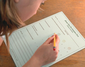A child is sitting at a wooden table, writing on a paper form with a pencil. The form contains questions and smiley face icons for feedback.