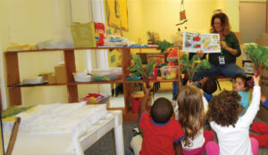 A teacher reads a story to a group of young children sitting on the floor, while they hold up green leaves resembling vegetables. The classroom has colorful decorations and shelves with various educational materials.